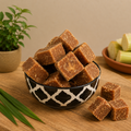 Bowl of brown square candies on a wooden table with a plant and green apples in the background.