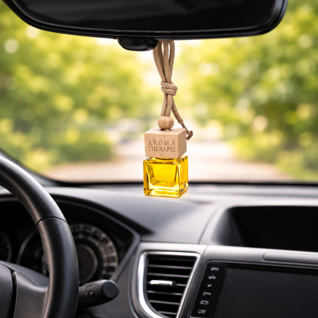 A car air freshener with a small square glass bottle filled with yellow liquid, a light wood cap engraved with 'AROMA THERAPIE', and a beige hanging cord, shown hanging in a car interior.