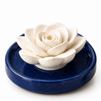 A white ceramic rose-shaped incense holder resting on a round navy blue saucer, set on a light beige marble surface.