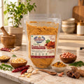 A stand-up pouch of Nut Sampoorn Signature Peanut Putana Instant Chutney filled with brownish-orange powder, displayed on a marble surface with peanuts, red chilies, garlic, and herbs around it.