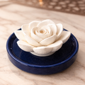 A white ceramic rose-shaped incense holder resting on a round navy blue saucer, set on a light beige marble surface.